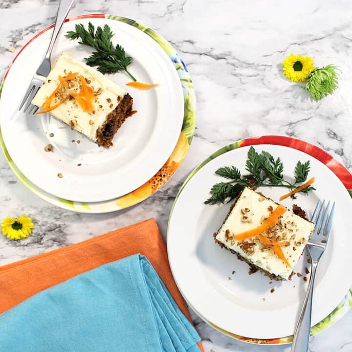 Overhead of two slices of carrot cake on white plates with orange and turquoise napkins.