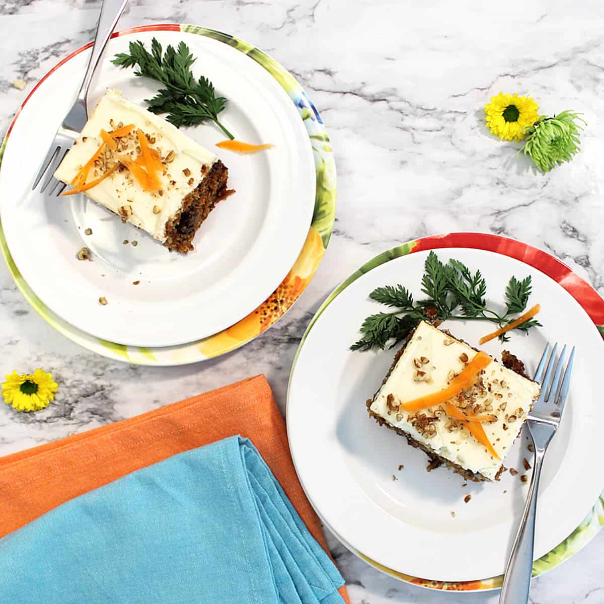 Overhead of two slices of carrot cake on white plates with orange and turquoise napkins.
