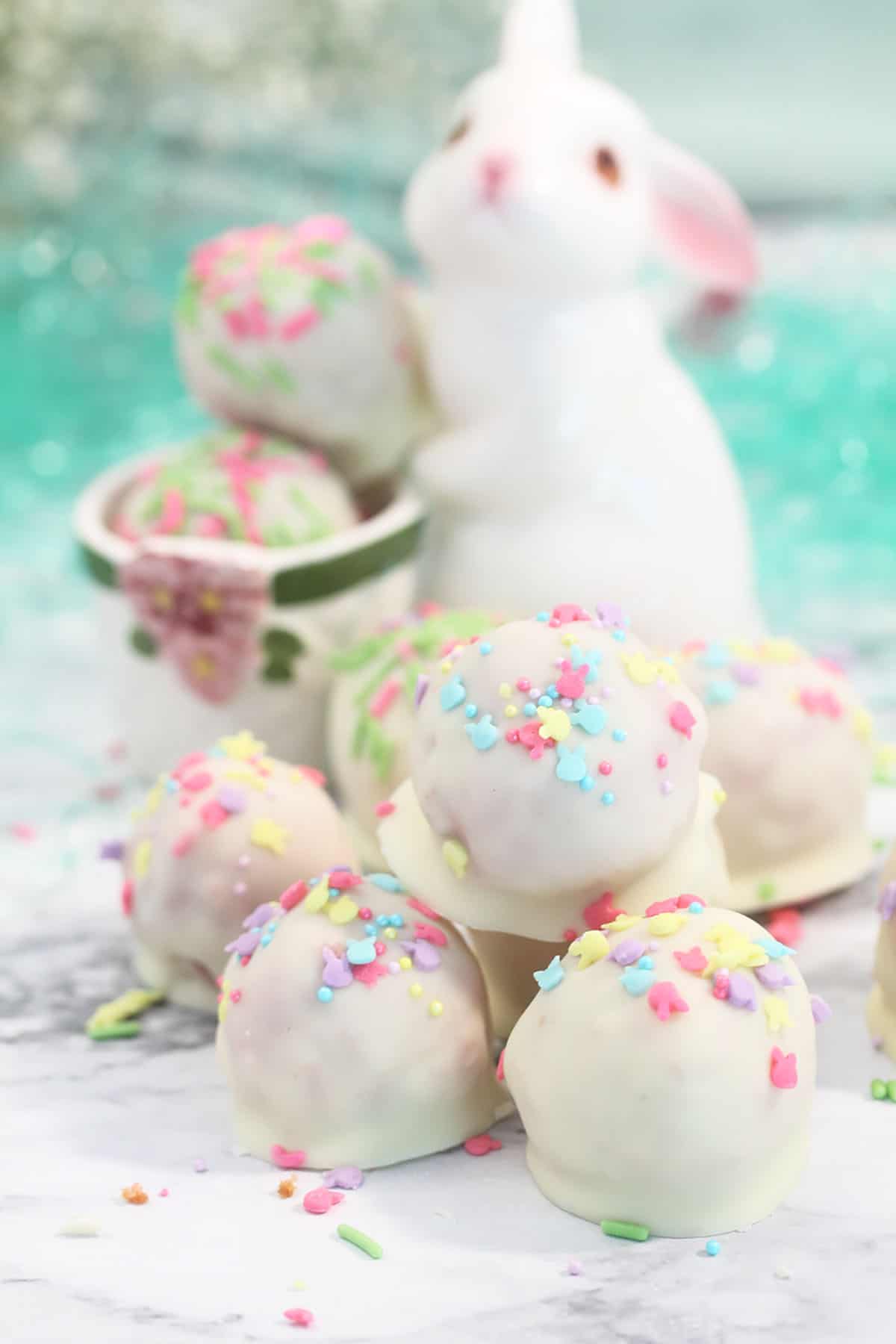 White ceramic bunny holding white chocolate coated cake balls on white table.