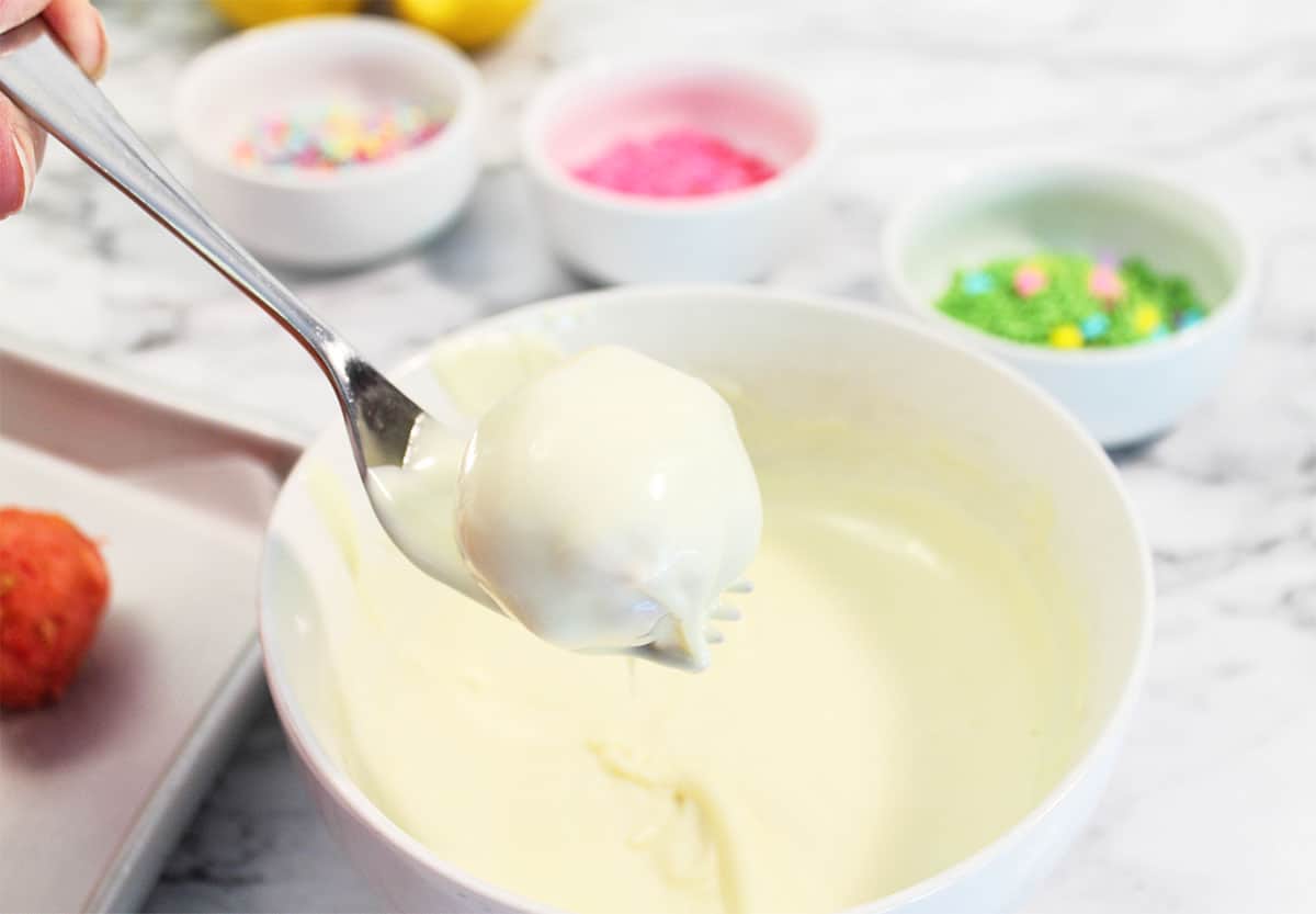 Dipping strawberry cake ball on fork in warm white chocolate.