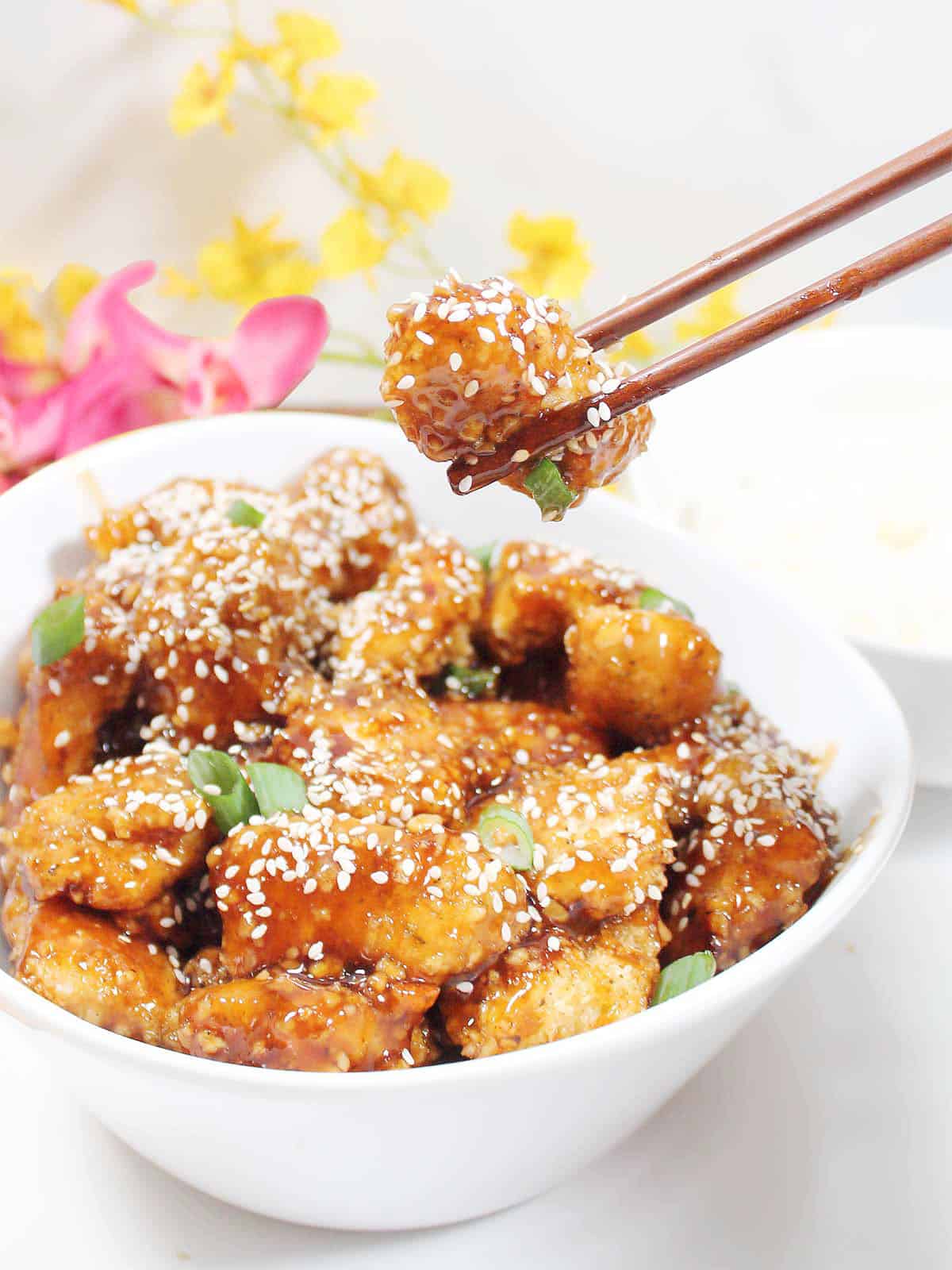 Holding piece of honey garlic chicken with chopsticks over serving bowl.