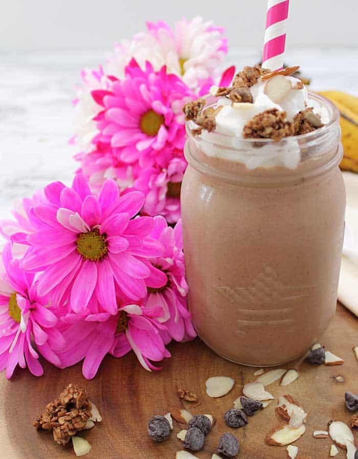 Healthy smoothie ready for breakfast , topped with whipped cream, with granola and almonds on top and flowers beside it.