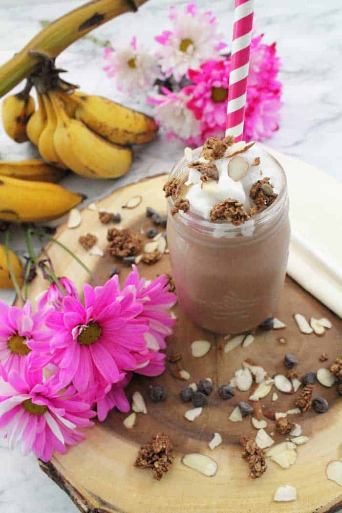 Topped smoothie in mason jar with red striped straw and bananas in back, pink flowers on side.