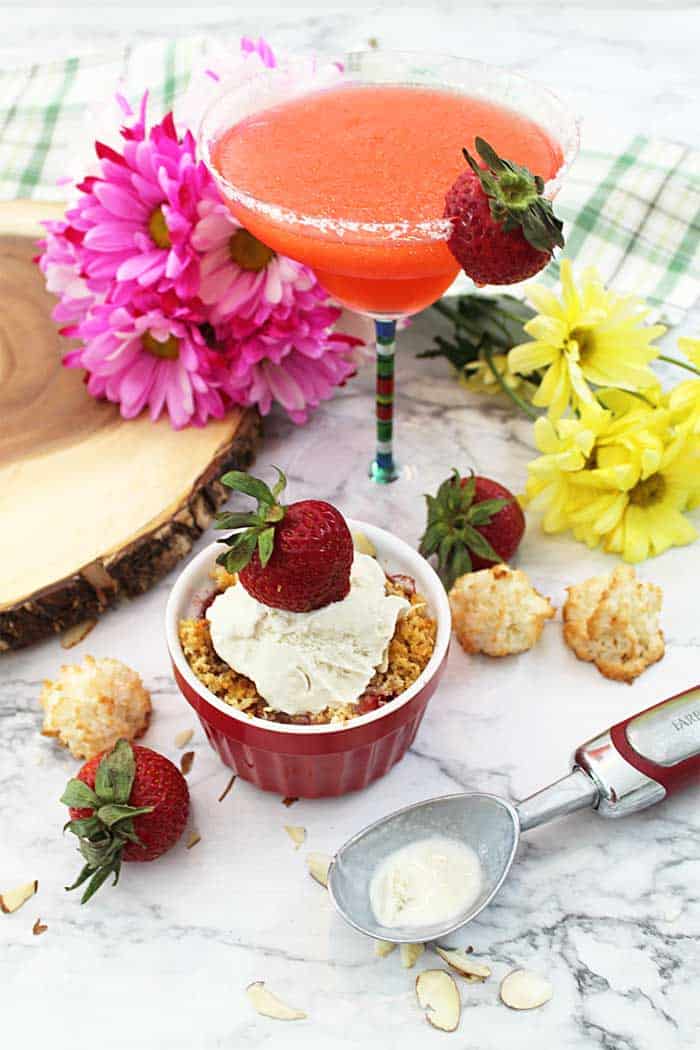 Strawberry Crisp on white tabe with margarita & flowers in background.