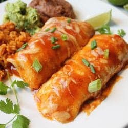 Closeup of enchiladas on white plate with lime slice rice guacamole and beans.