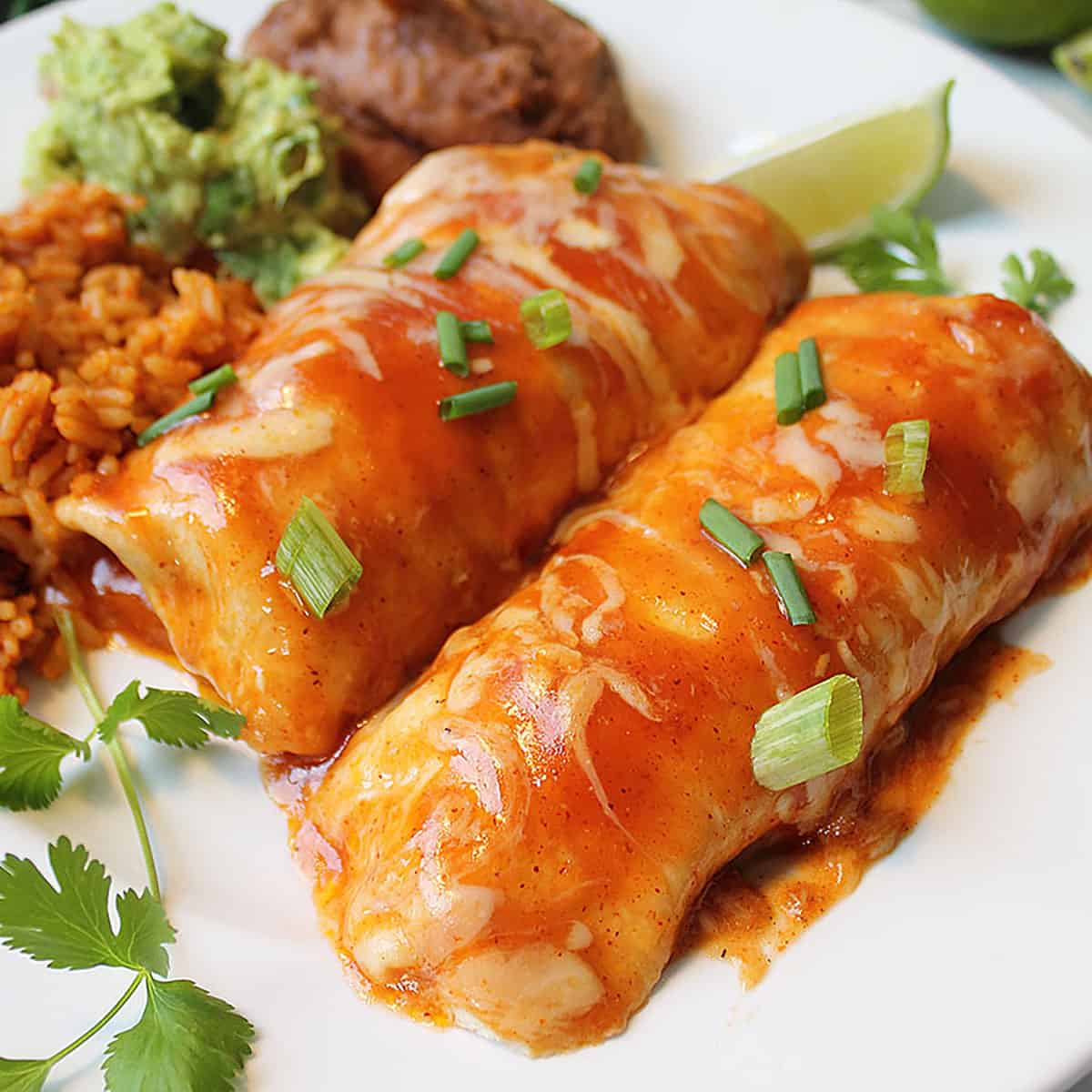 Closeup of enchiladas on white plate with lime slice rice guacamole and beans.
