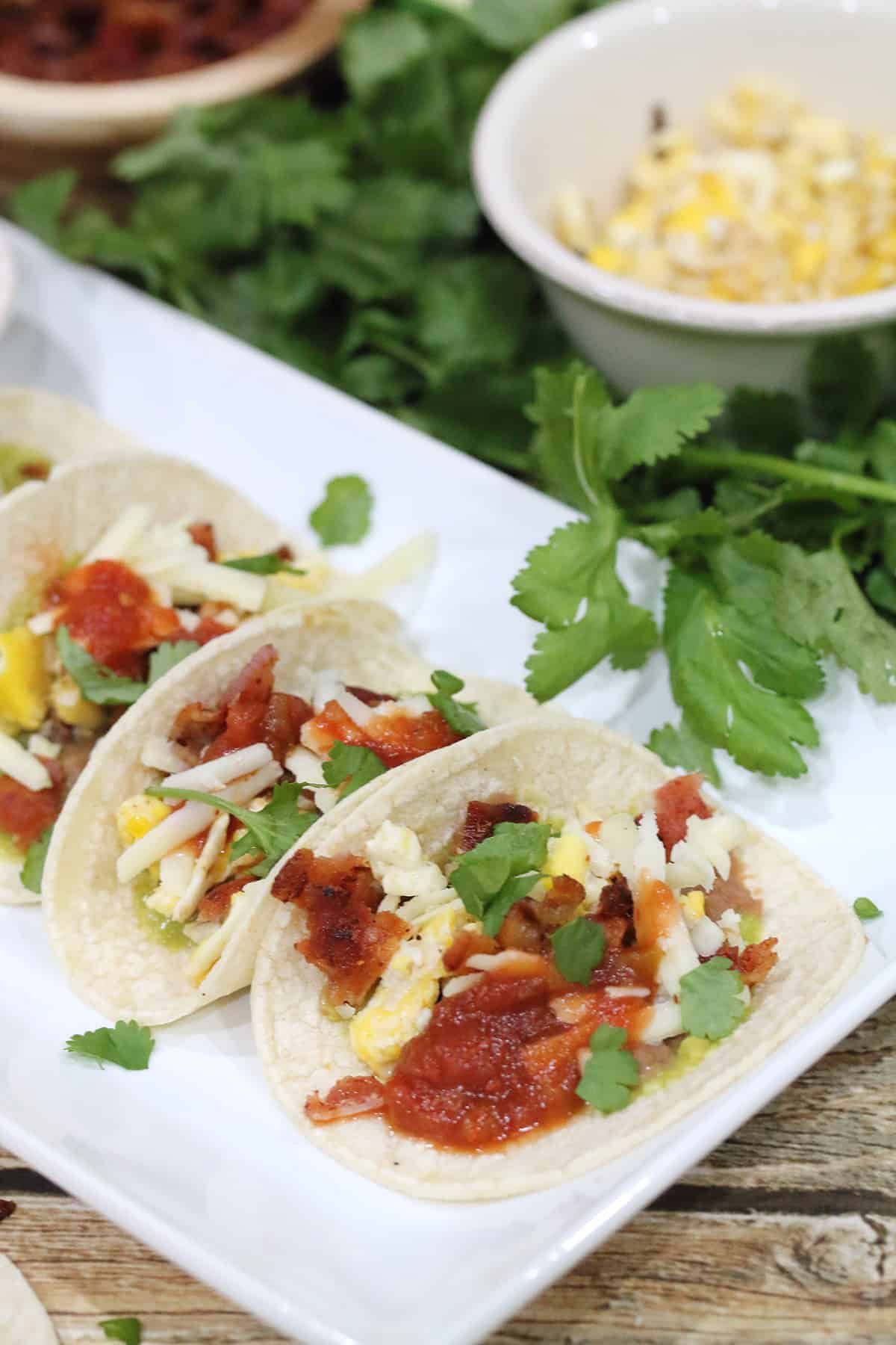 Plated Mexican breakfast tacos with cheese and bacon bowls in back.