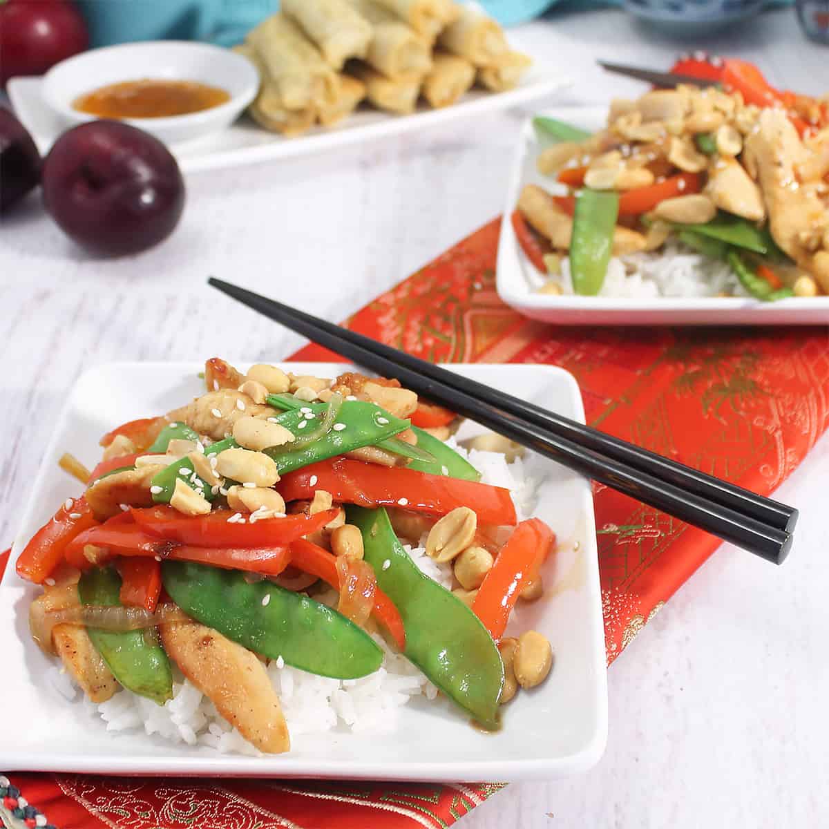 Two plated servings of chicken stir fry with plum sauce with egg rolls in background.