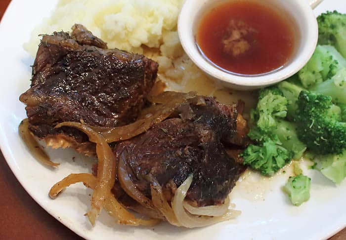 Closeup of short ribs on white plate next to mashed potatoes and broccoli.