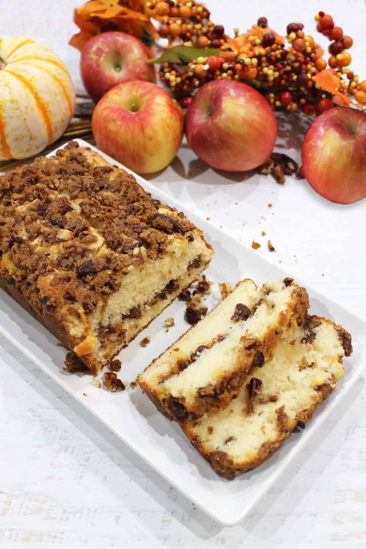 Cinnamon Apple Streusel Bread sliced on white platter with fall apples and pumpkin in back.