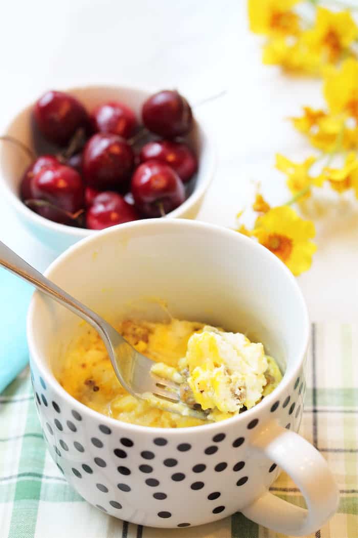 Spoonful of breakfast scramble in mug with cherries and yellow flowers in background.