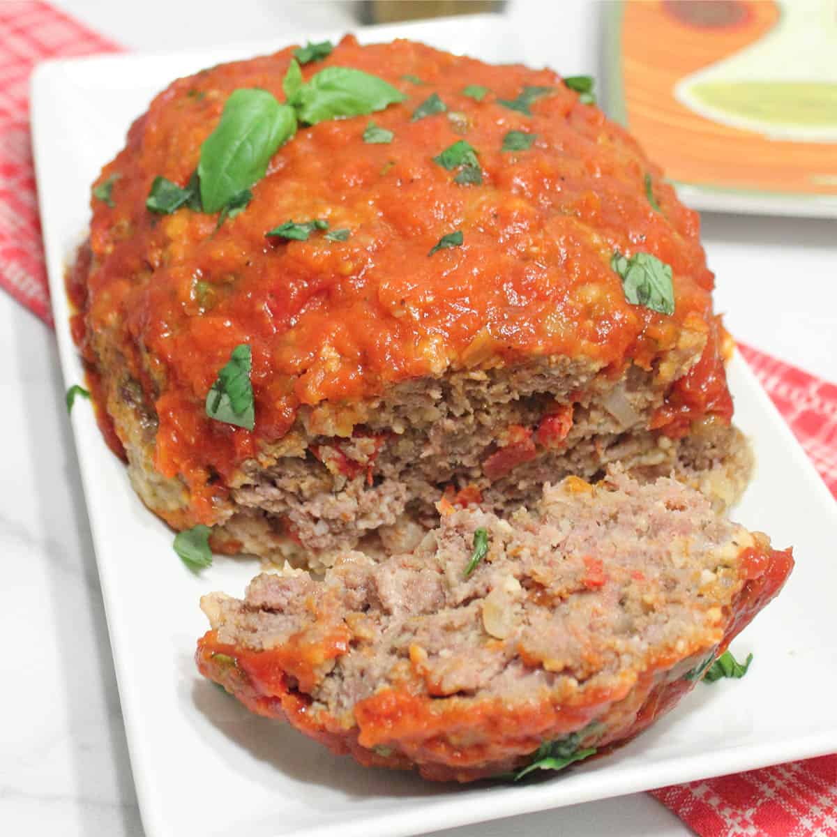 Meatloaf on serving plate with one slice and basil garnish.