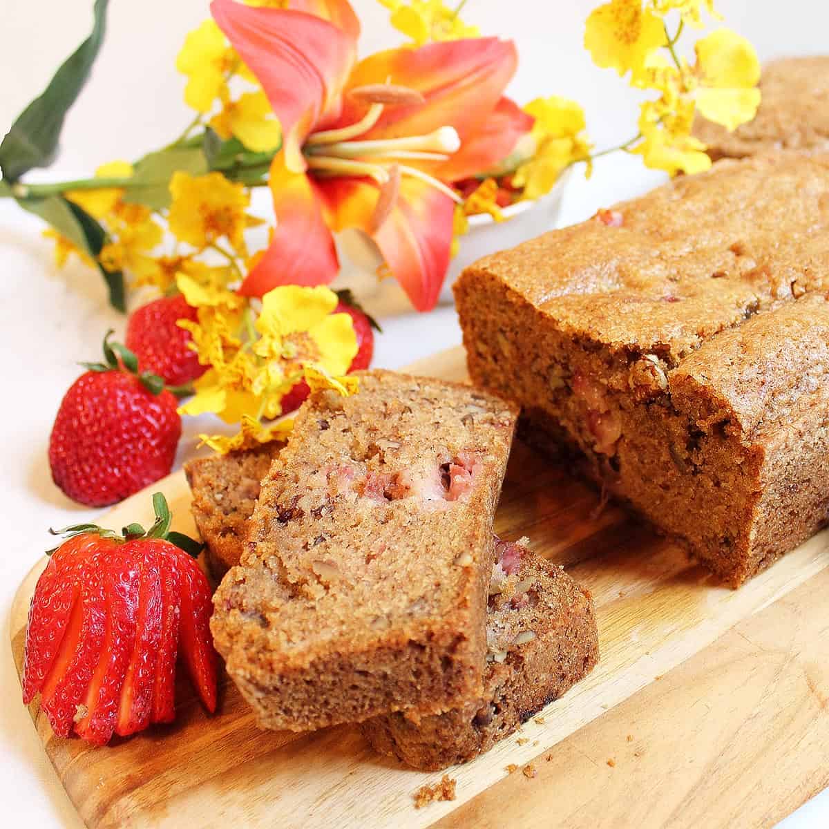 Closeup of strawberry quick bread on cutting board with strawberries and full loaf in background.