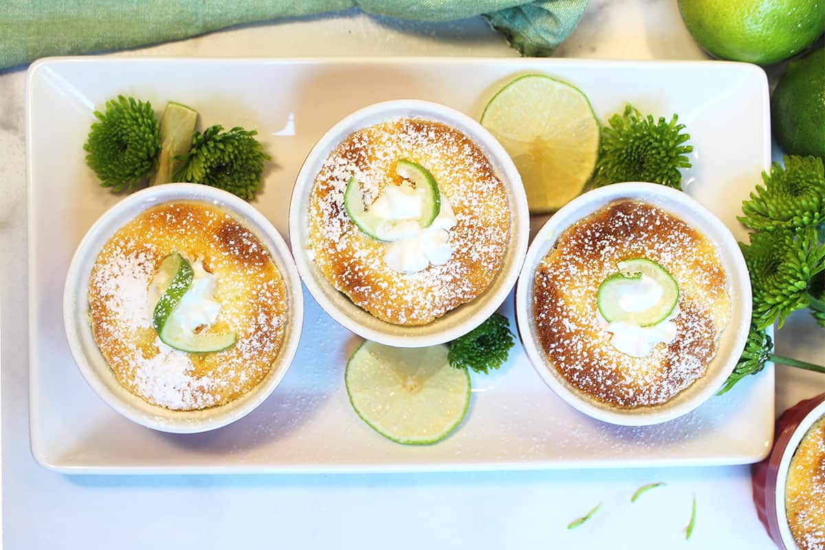Overhead of 3 key lime cake ramekins on white platter with green flowers and lime slices.