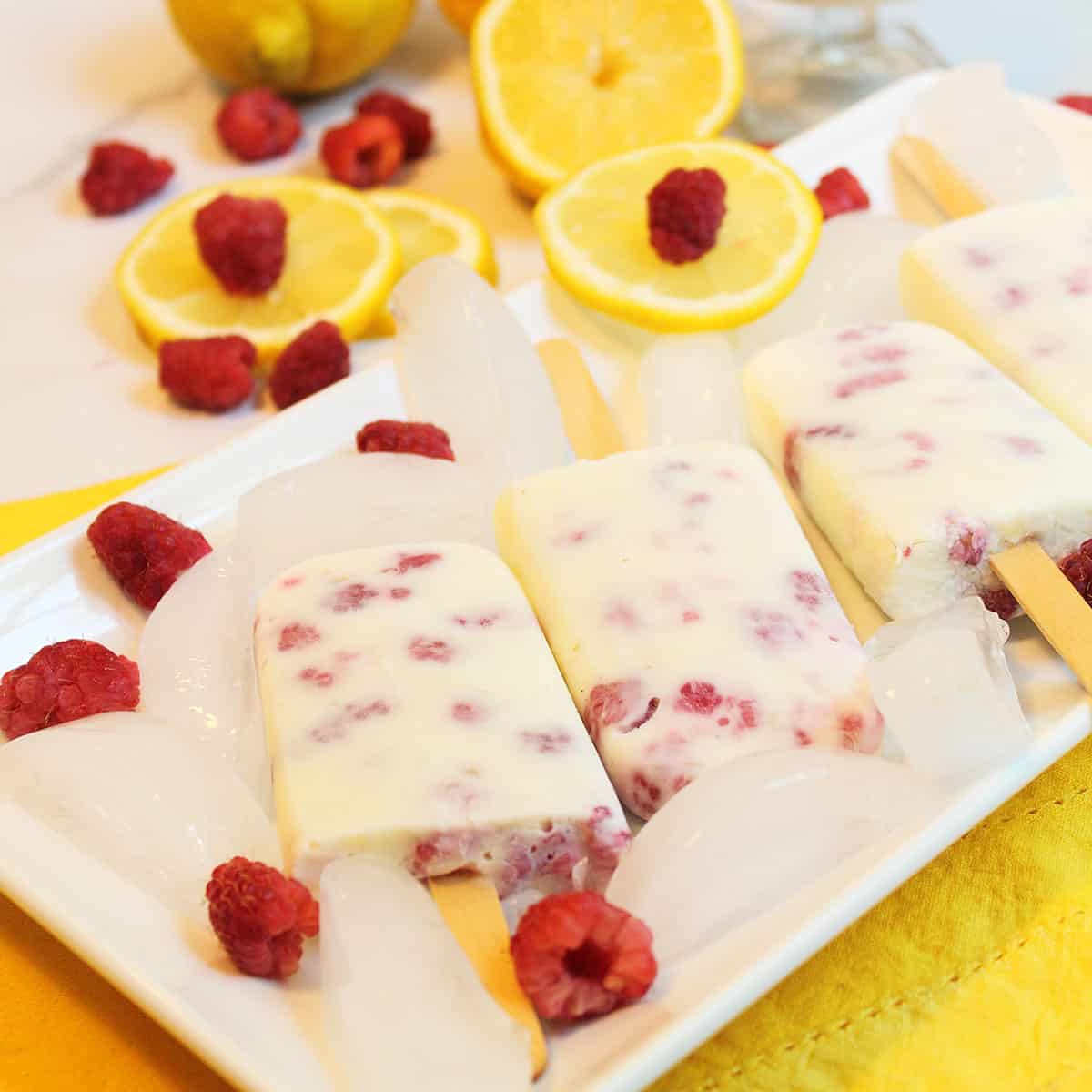 Popsicles on white platter with ice cubes and lemons and yogurt in serving cup behind it.