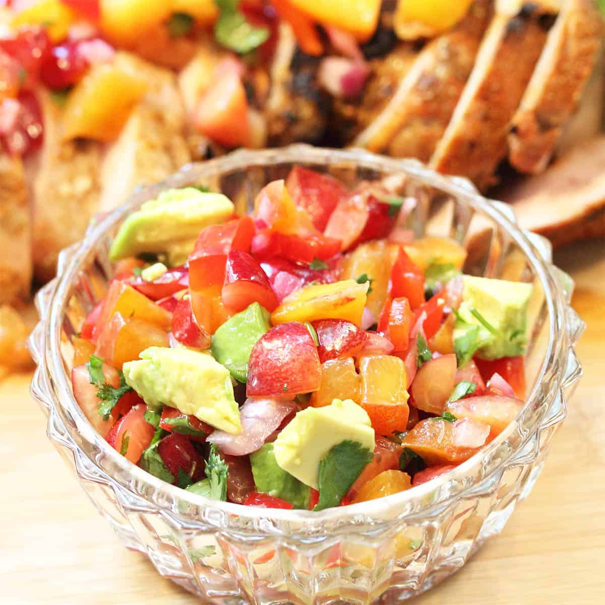 Closeup of peach cherry salsa tossed with avocado in glass bowl with pork in background.