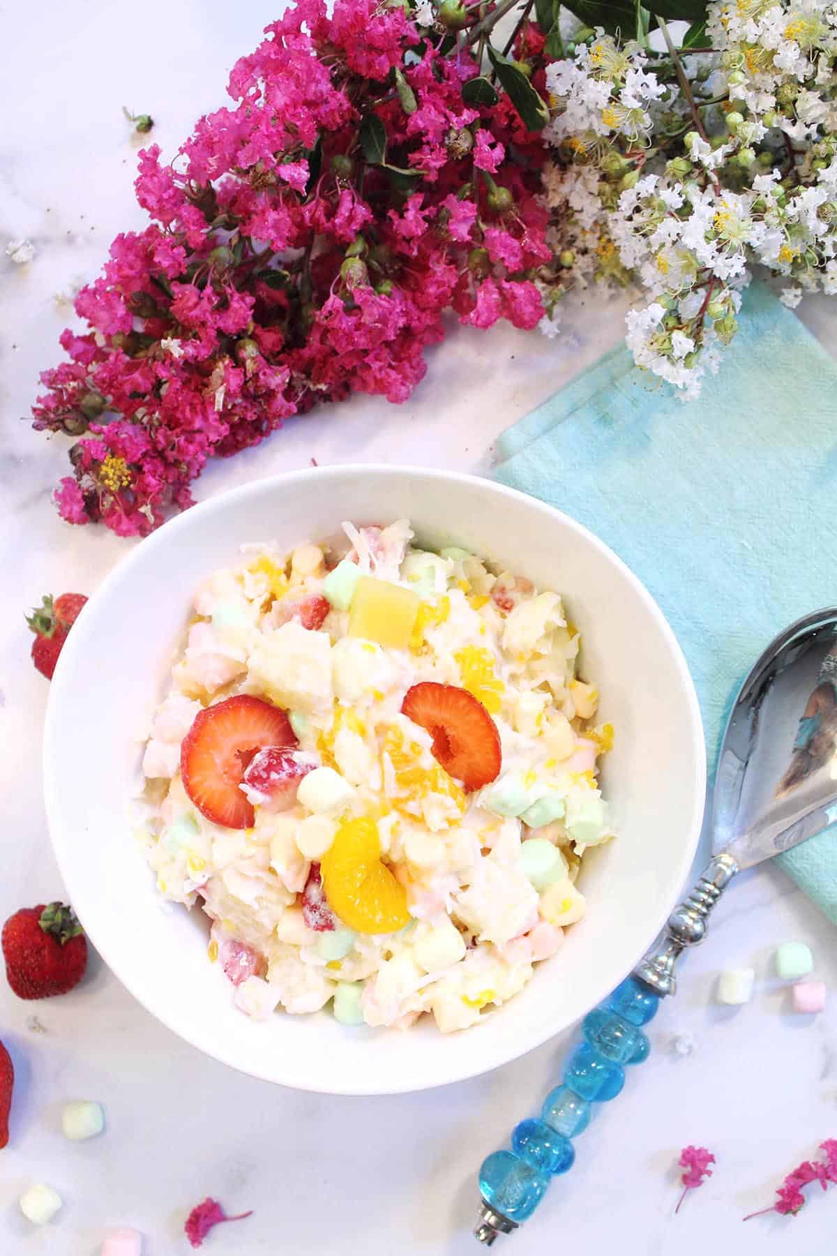 Overhead of white bowl of ambrosia salad covered with yogurt with serving spoon, green napkin & flowers on side.