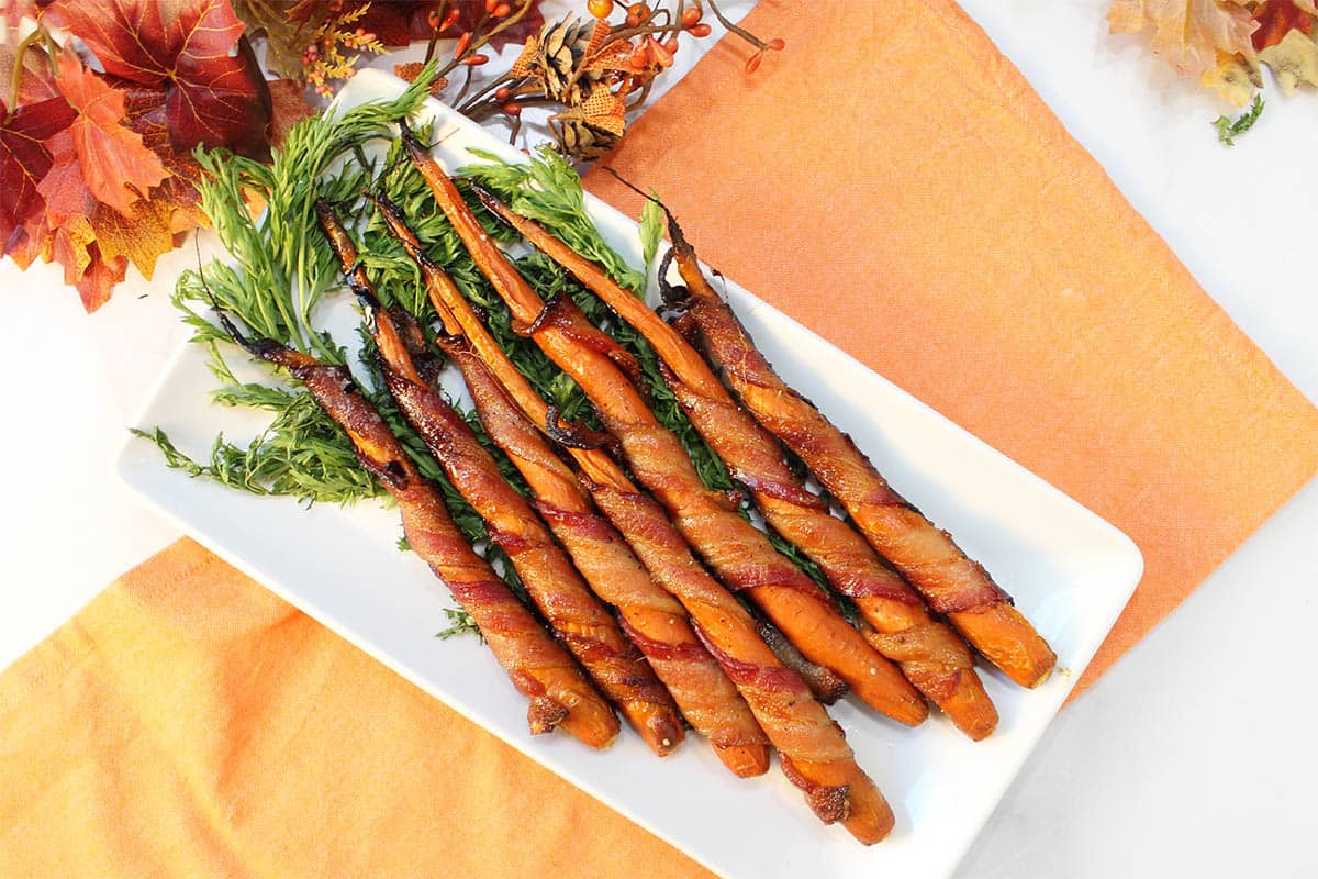 Maple Bacon Wrapped Carrots on white platter with carrot greens on orange napkin.