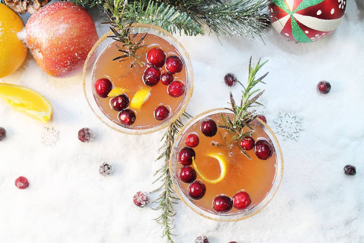 Overhead of vodka apple cider cocktails on snow with orange apple and ornament around them.