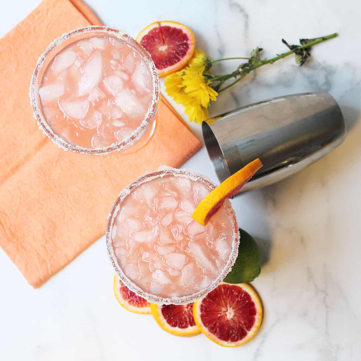 Overhead of two cocktails surrounded by blood orange slices orange napkin and shaker.