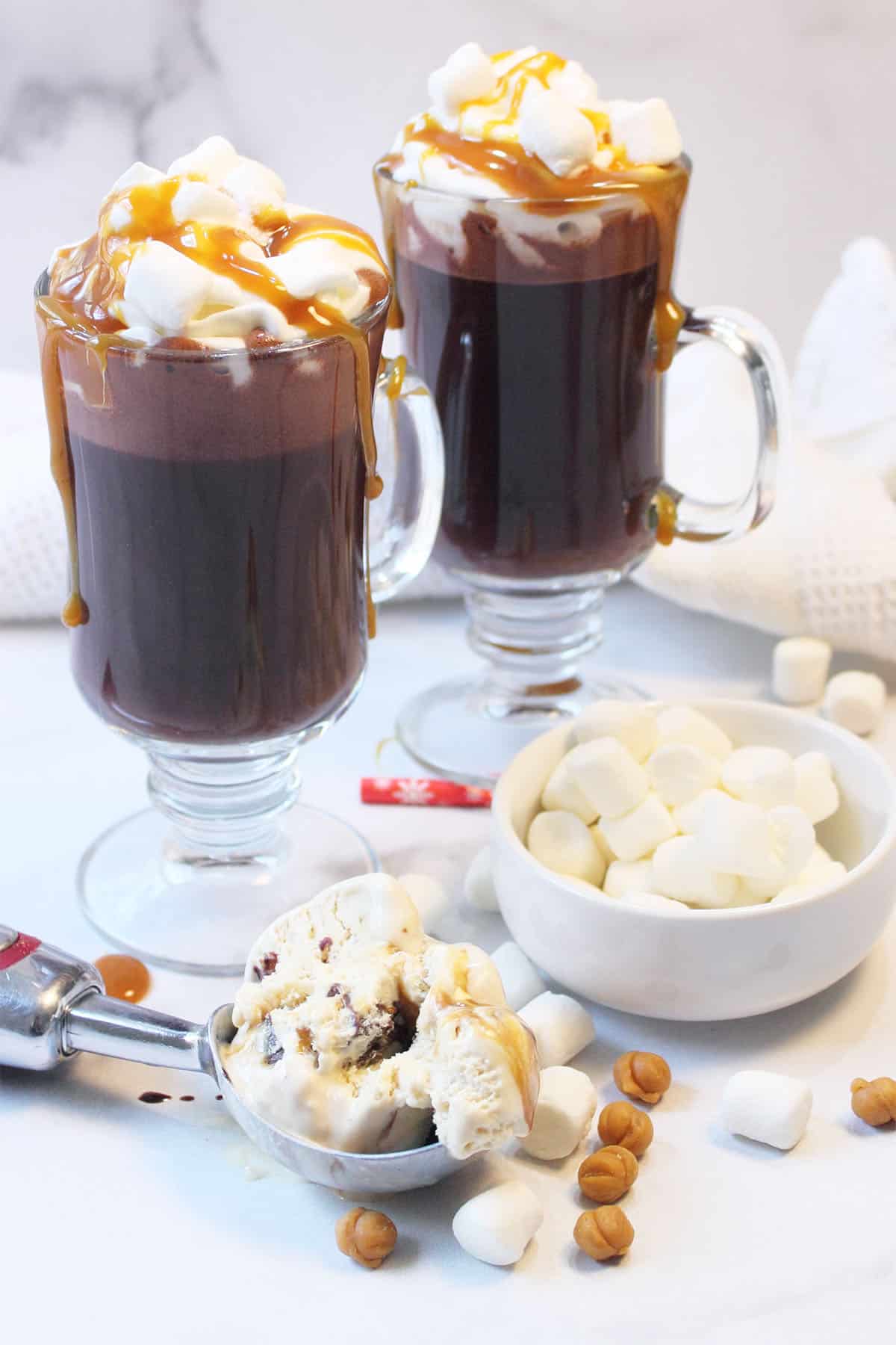 Two hot chocolates with whipped cream and caramel sauce with scoop of ice cream and marshmallows on the table.