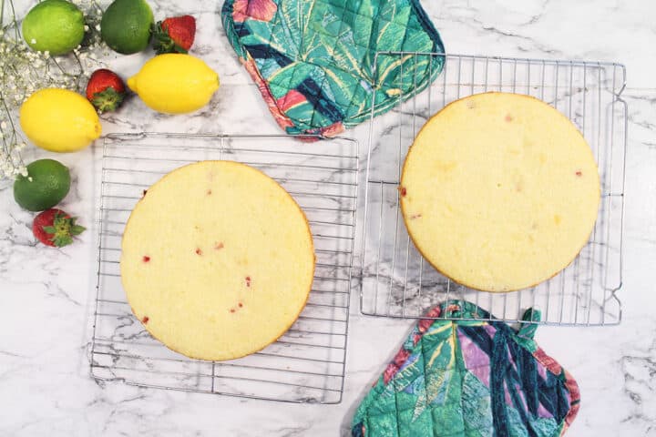 Cake layers cooling on metal racks with fruit and colorful pot holders around them.