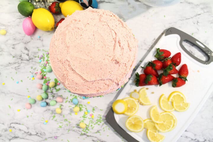 Frosted cake on cake stand ready to decorate. Fruit on cutting board next to cake.