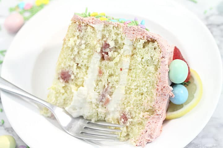 Closeup of slice of lemon cake on white plate with pastel candy and lemon slice garnish.