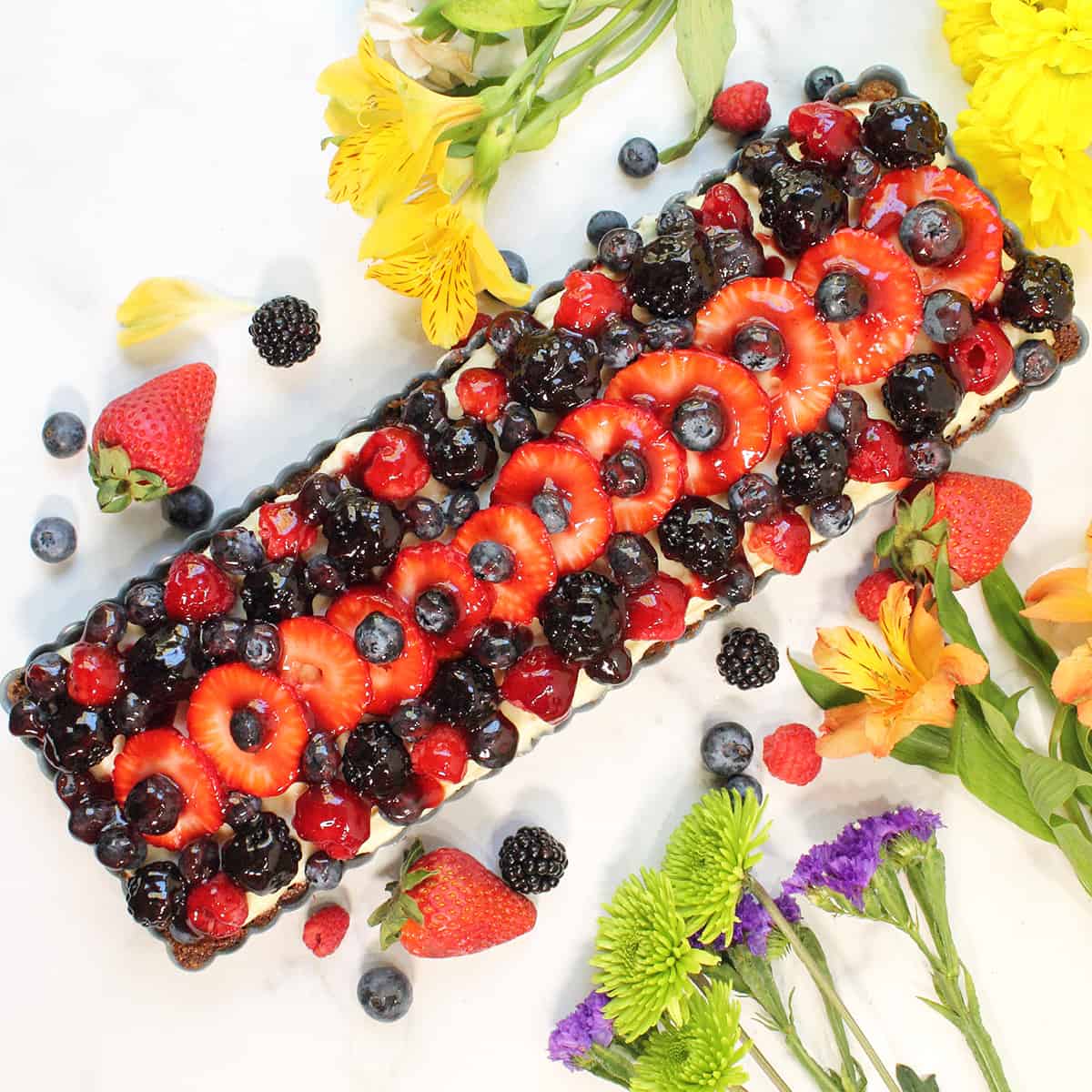 Fresh Fruit Tart on marble table with flowers and fruit around it.