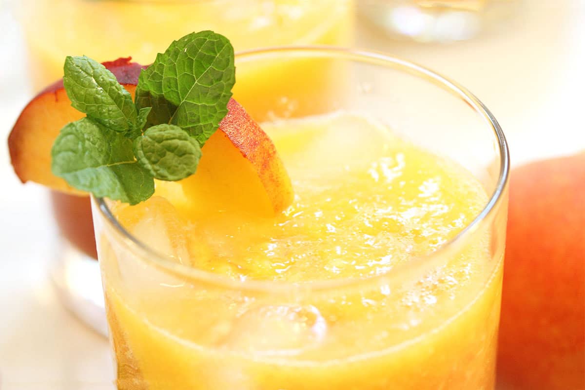 Closeup of top of glass filled with peach drink and garnished with peach slice and mint.