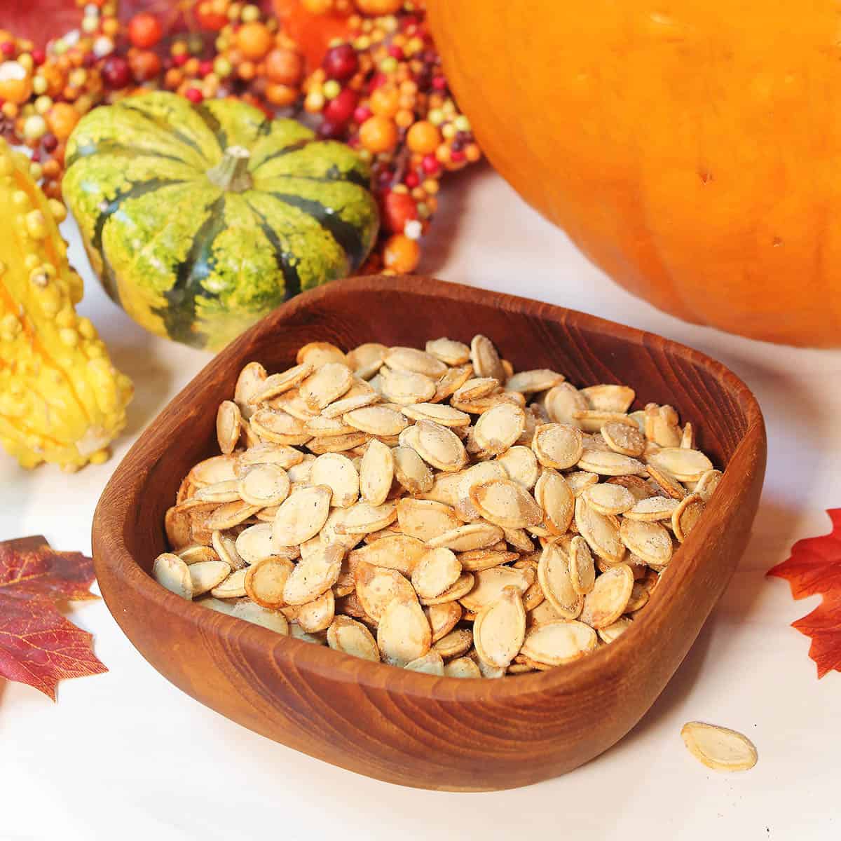 Wooden bowl filled with seeds on white table with gourds and fall leaves.