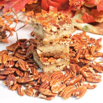 Stacked pecan pie bars with shortbread crust amidst pecans.