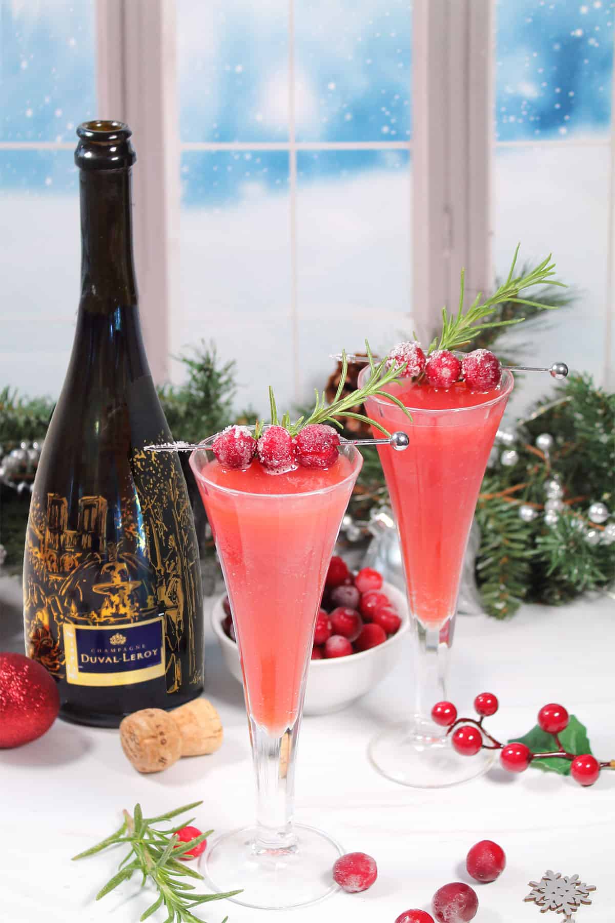 Two cranberry mimosas in flutes with champagne bottle by window.