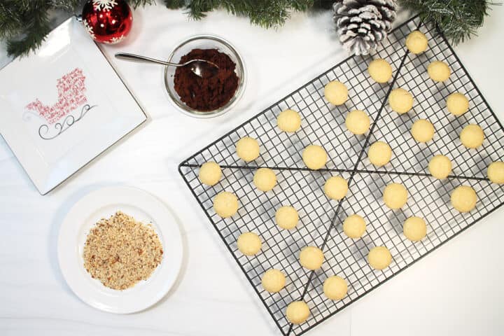 Baked cookies on cookie sheet next to chocolate frosting and chopped nuts on white table..