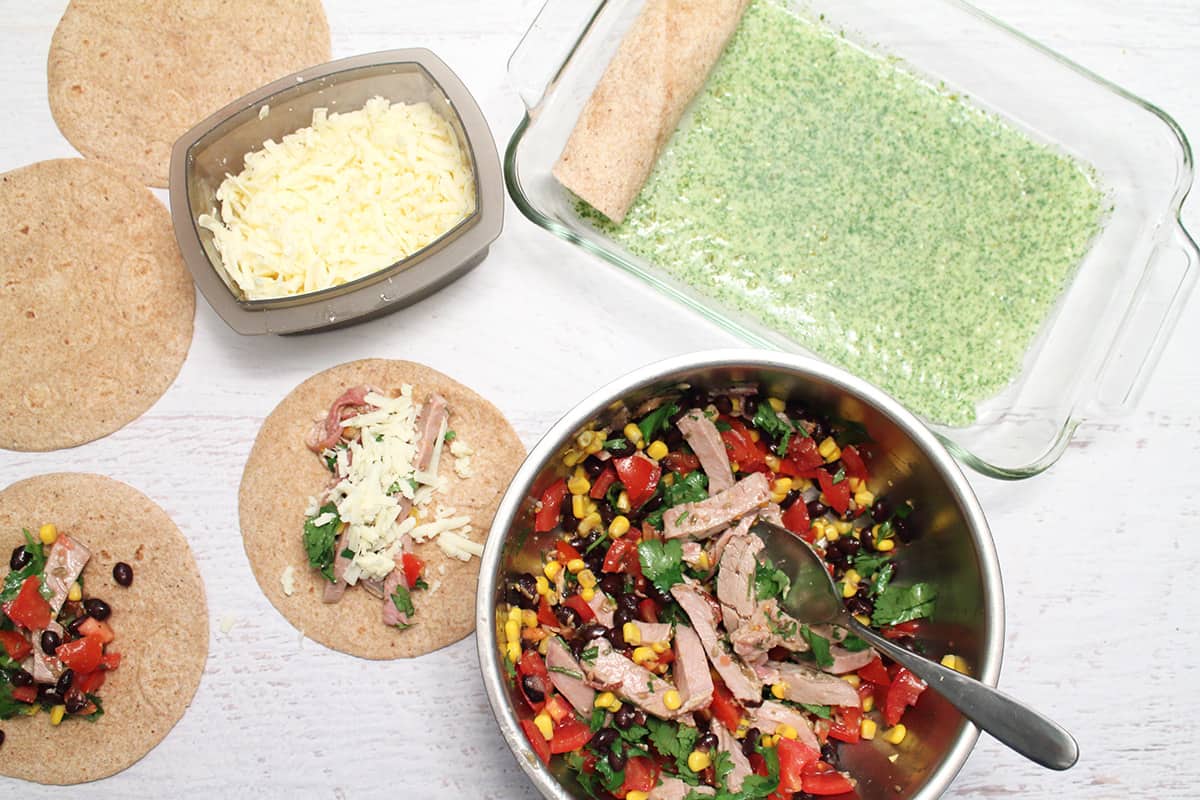 Assembly line to make enchiladas with green sauce in bottom of dish and tortillas with filling.