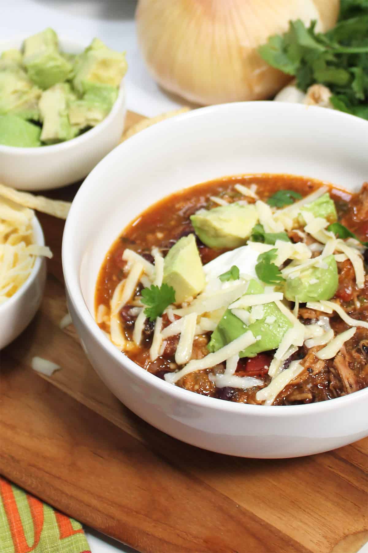 Bowl of chili on wooden board with cilantro, onion and chopped avocado.