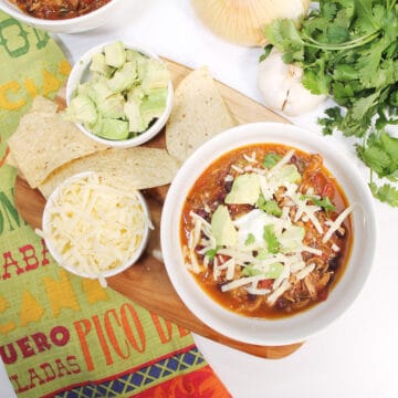Garnished stew in white bowl on wooden board with chips and sides.