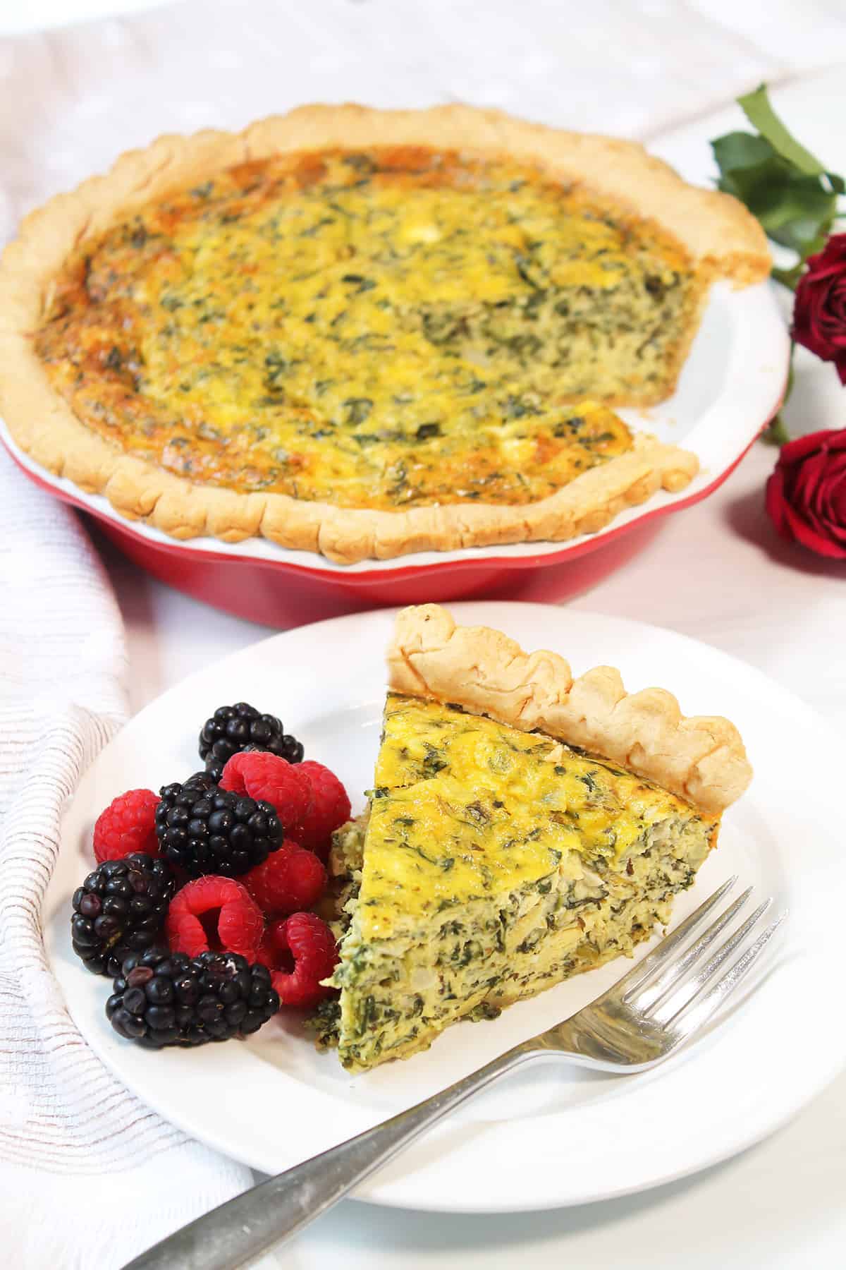 Slice of spinach artichoke quiche on white plate with whole quiche in back.