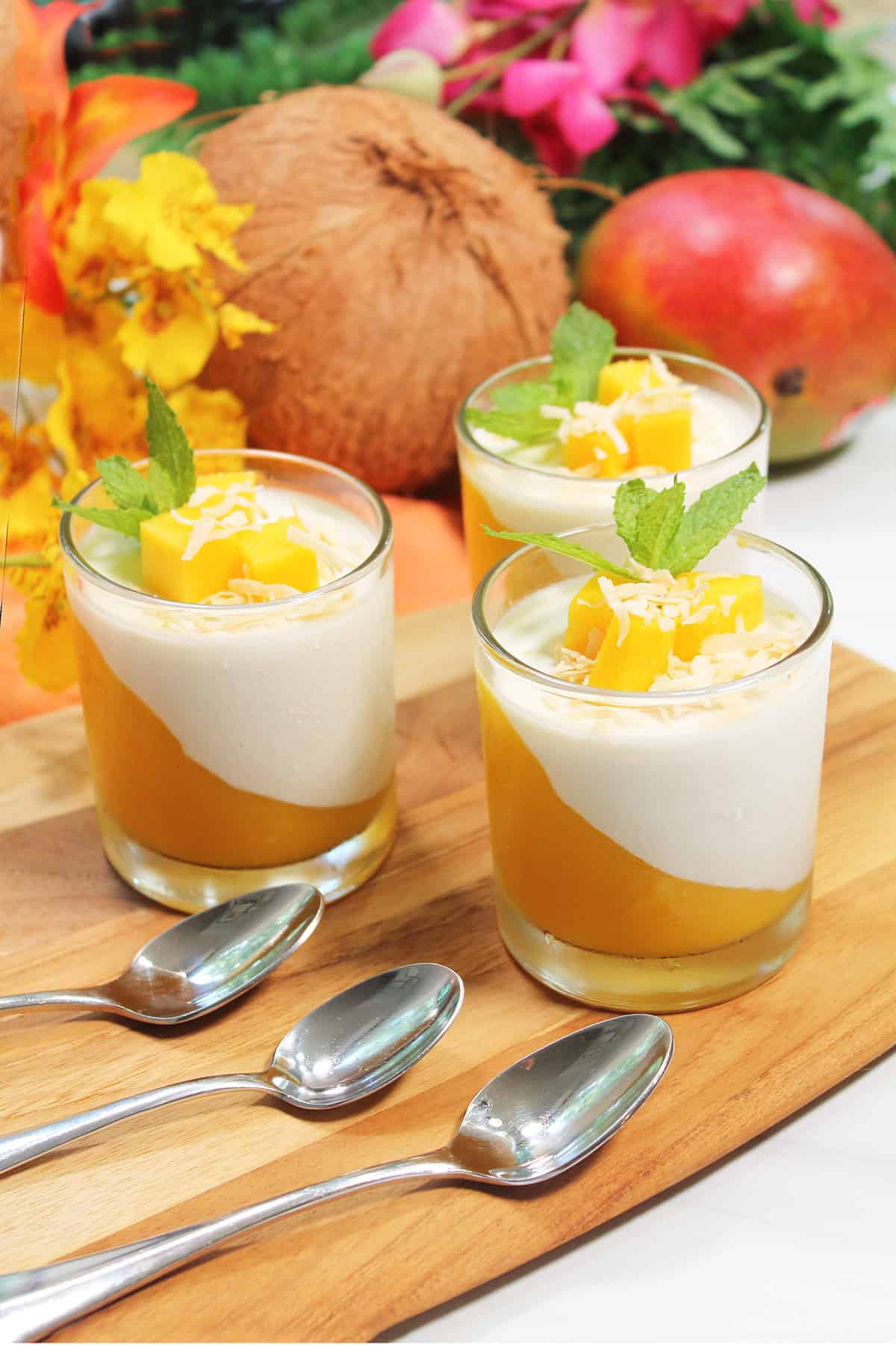 Three mango panna cotta dishes on wooden board with spoons.