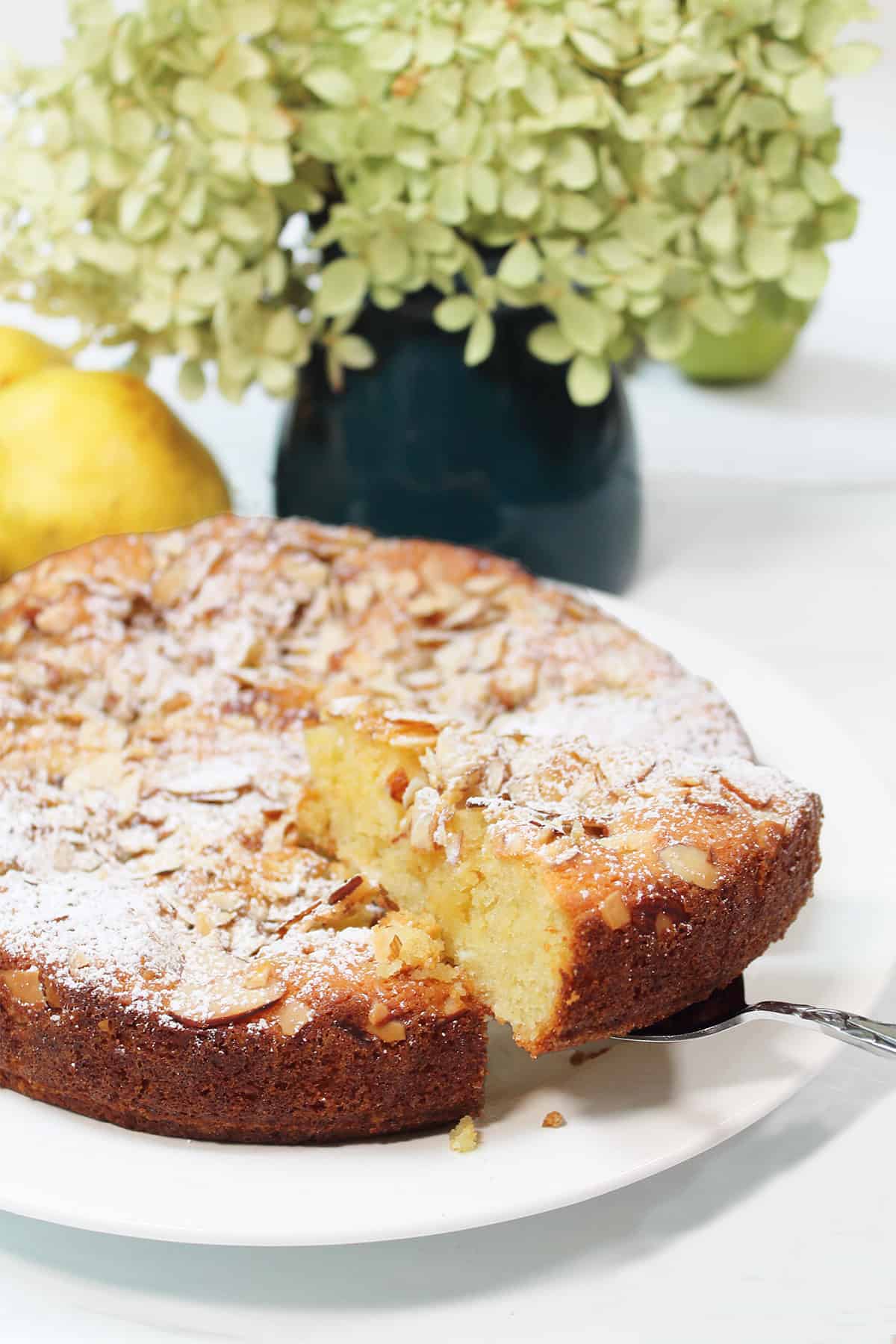 Lifting slice of almond cake with flowers in back.