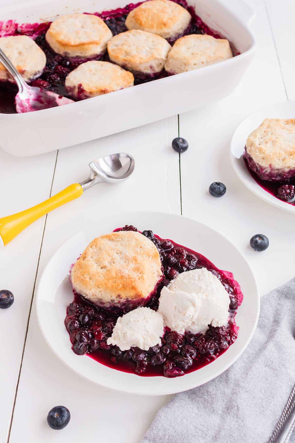 Mixed berry cobbler serving with ice cream in front of baking dish.