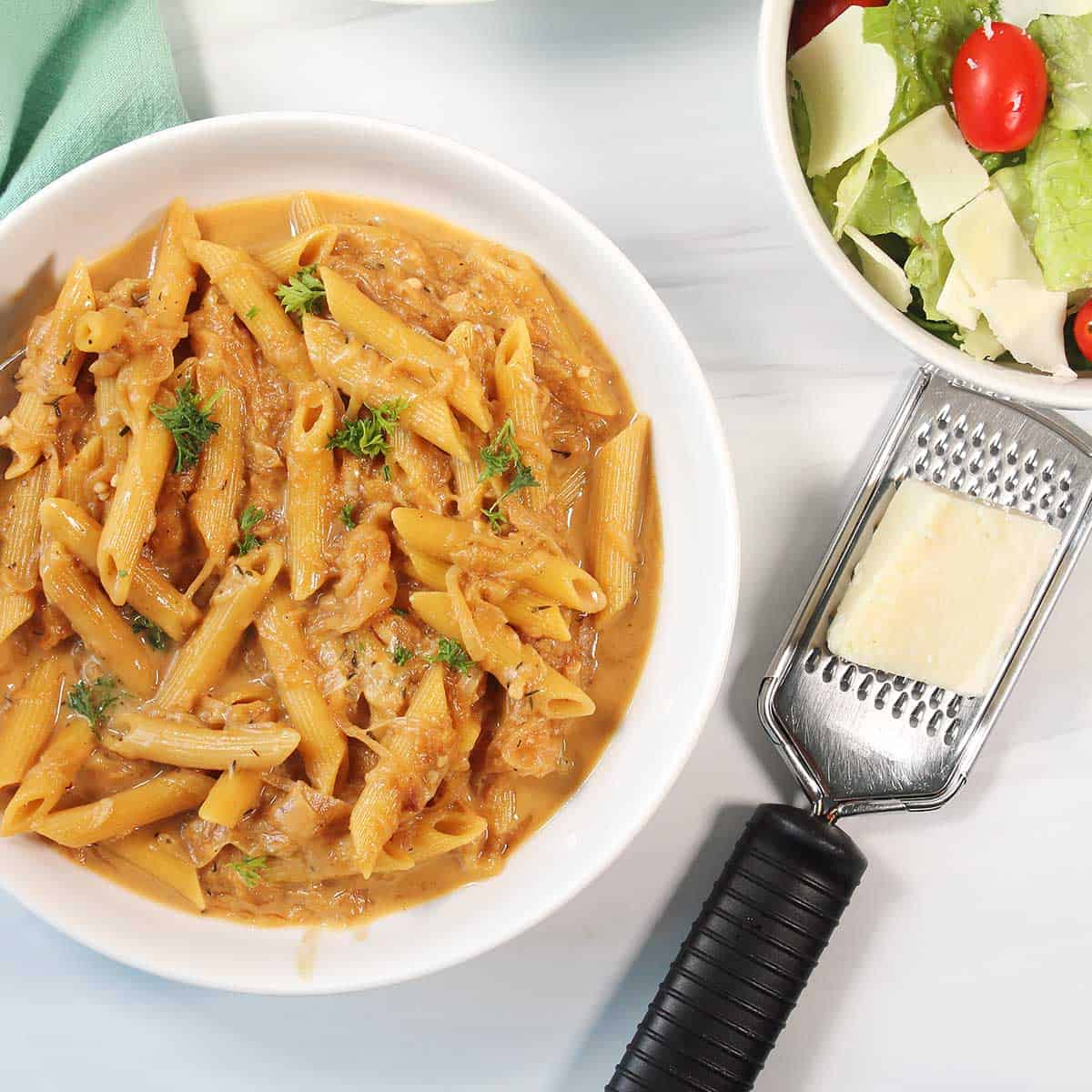 Overhead of French Onion Pasta in white serving bowl with Parmesan cheese and salad.