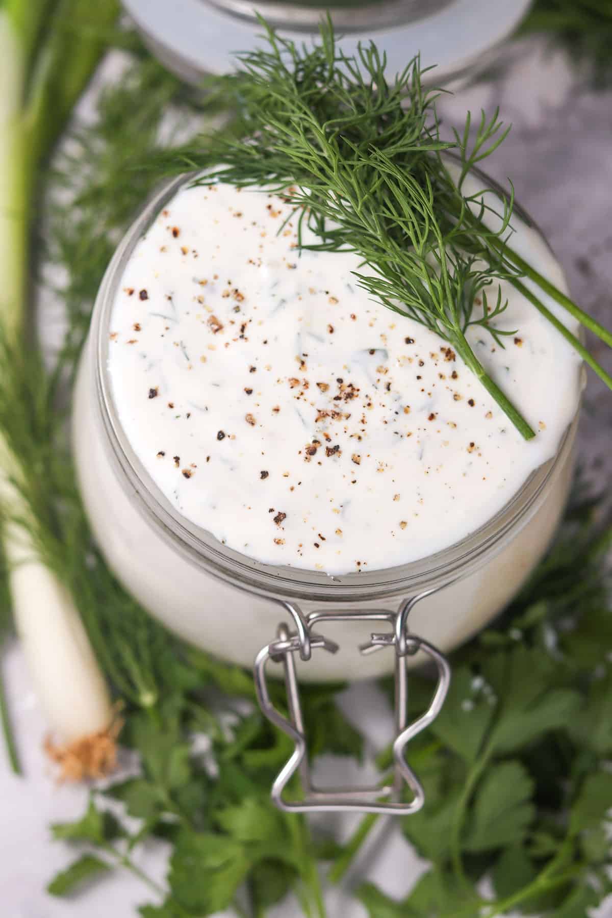 Creamy ranch dressing in jar surrounded by herbs.