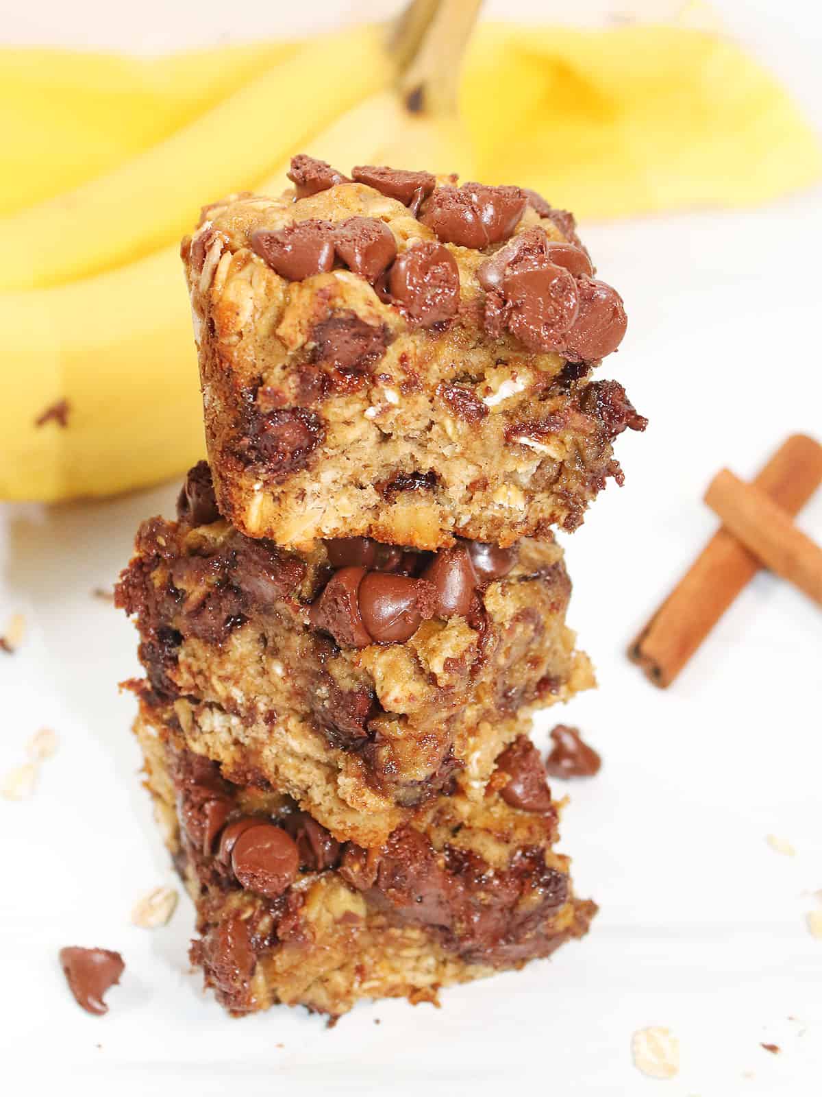 Three banana oatmeal chocolate chip bars stacked on table with bananas.