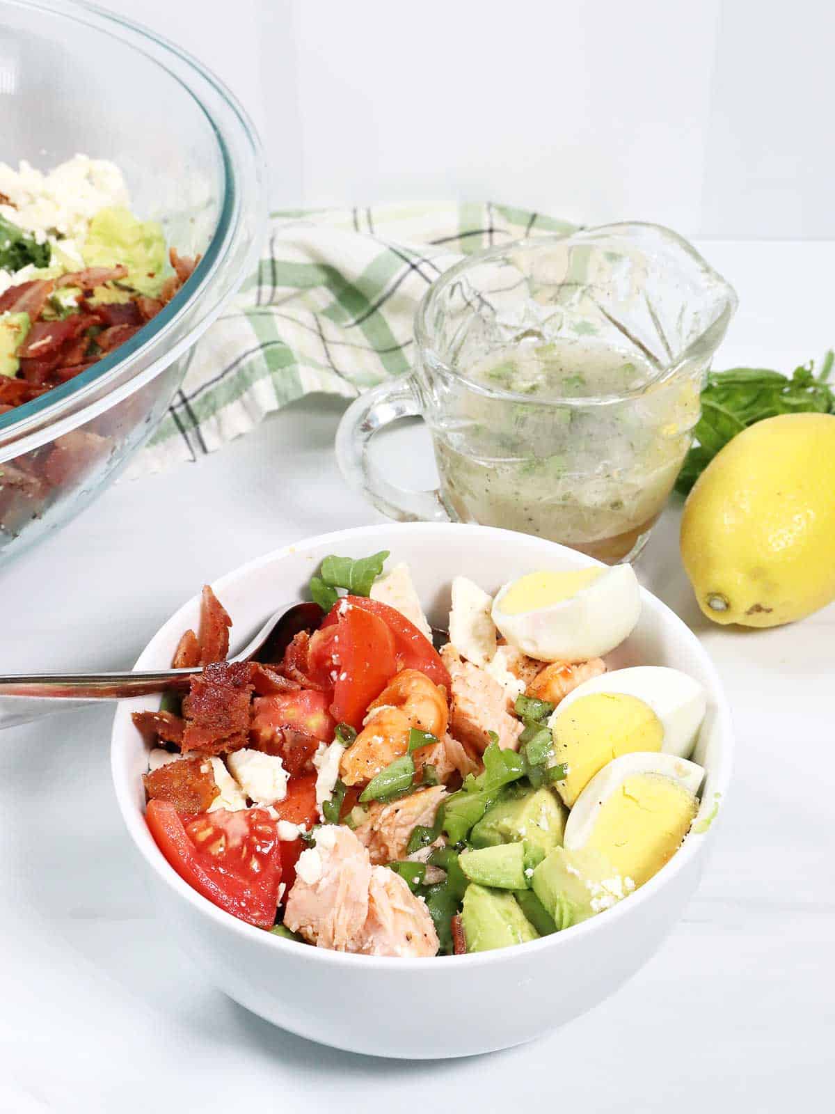 A serving of salmon cobb salad in front of large salad bowl.